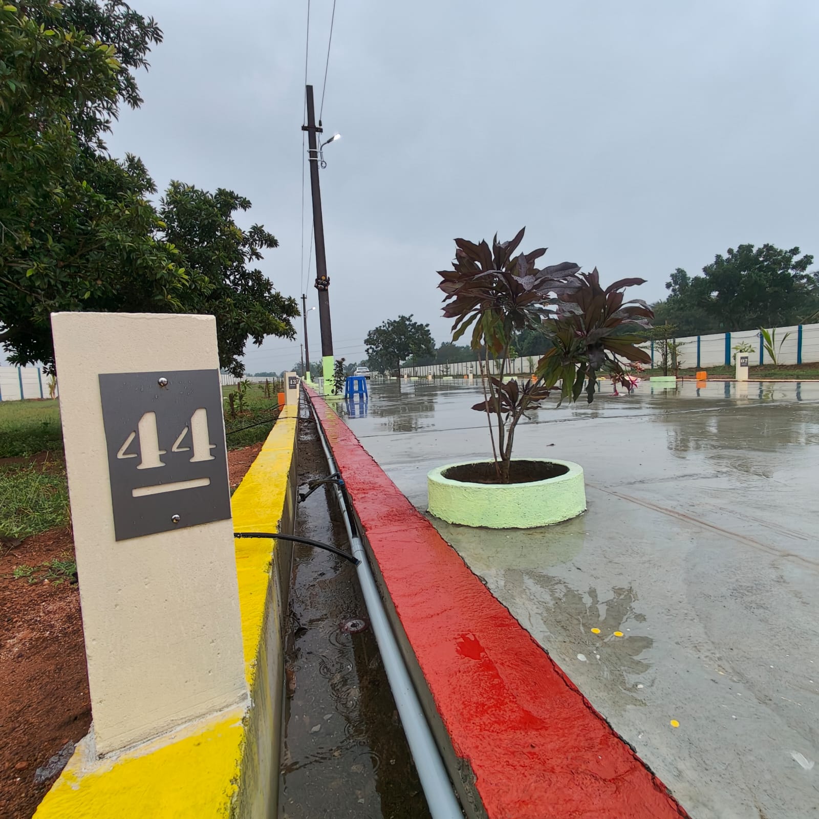 Plot number 44 at RK Happy Nest residential layout with paved road, drainage, street lights, boundary wall, and landscaped plantation area after rain.